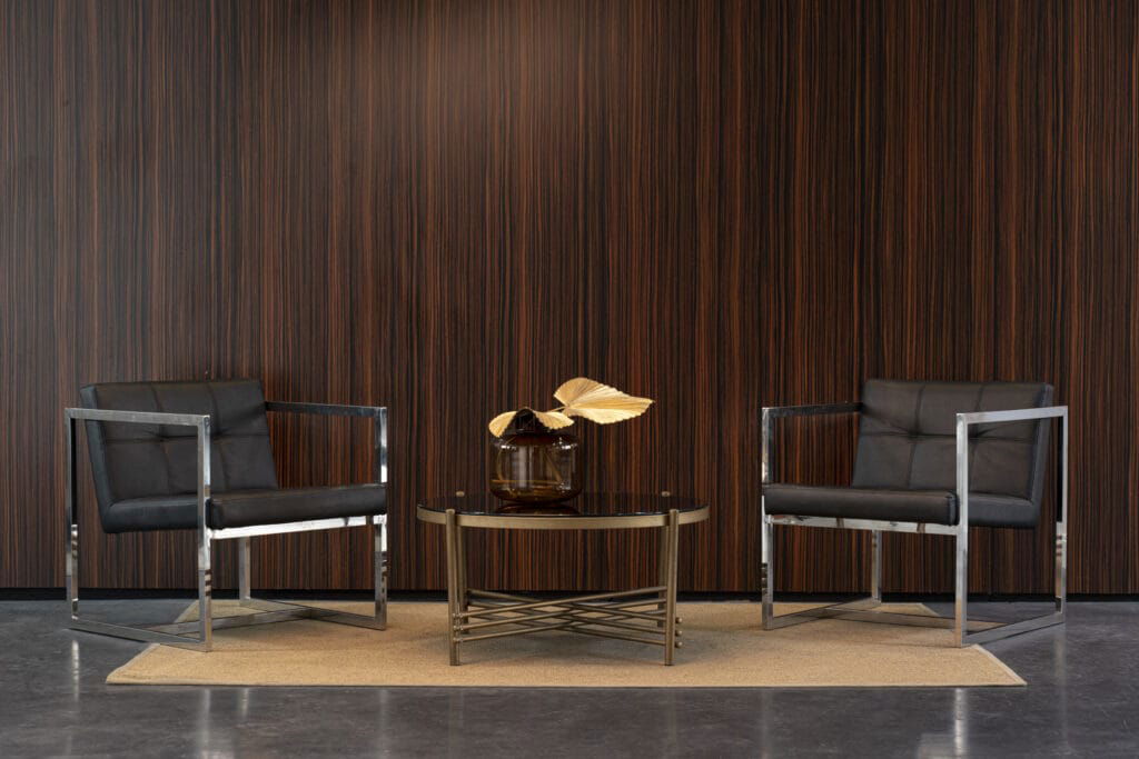 Two modern black armchairs face each other, separated by a glass coffee table with a decorative gold-leaf centerpiece. Set against a dark wooden wall installed by a top-notch wallpaper contractor, they rest on a beige rug.